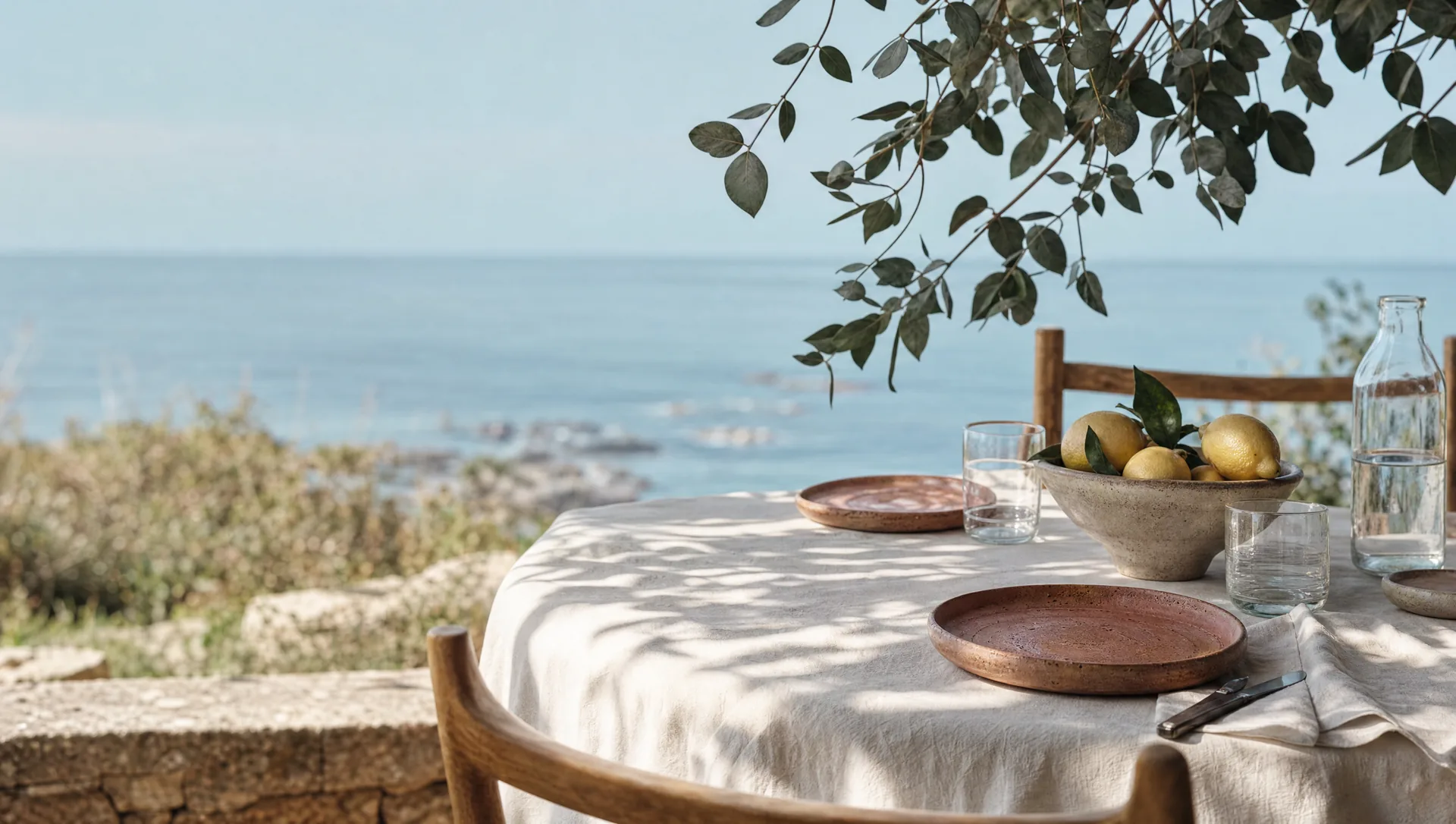 A linen-covered lunch table under a tree above the sea, a bowl of lemons at its centre